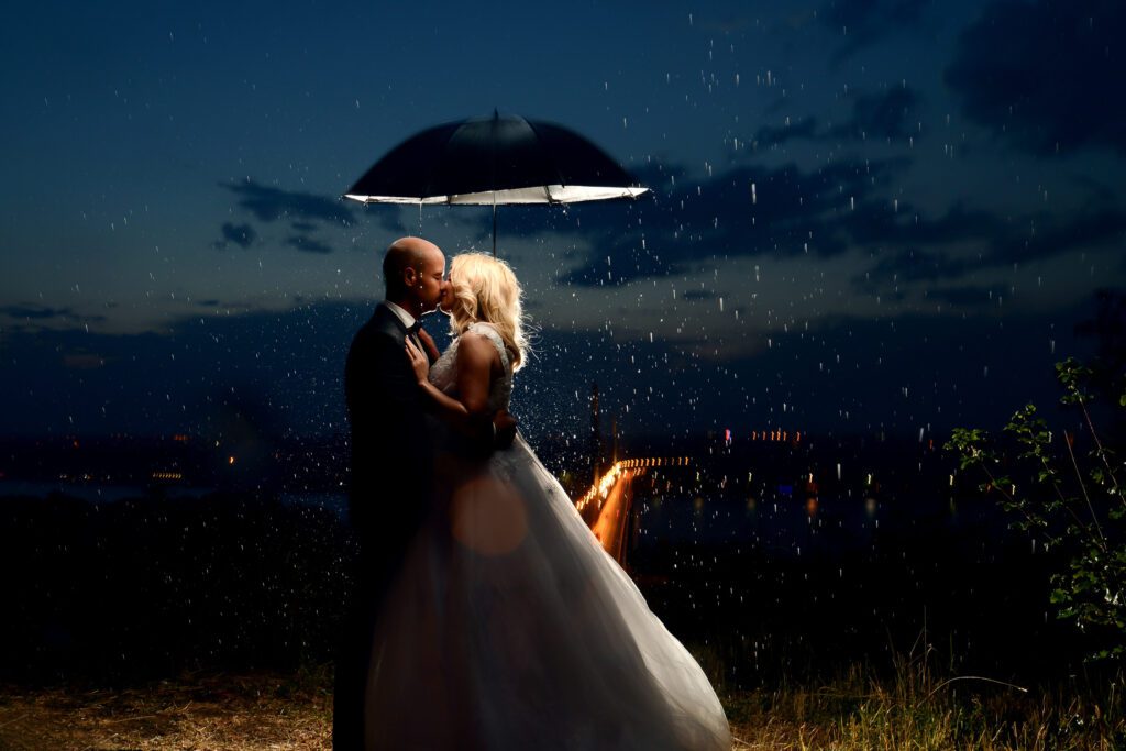Newlyweds kiss under an umbrella on a rainy wedding day at night.